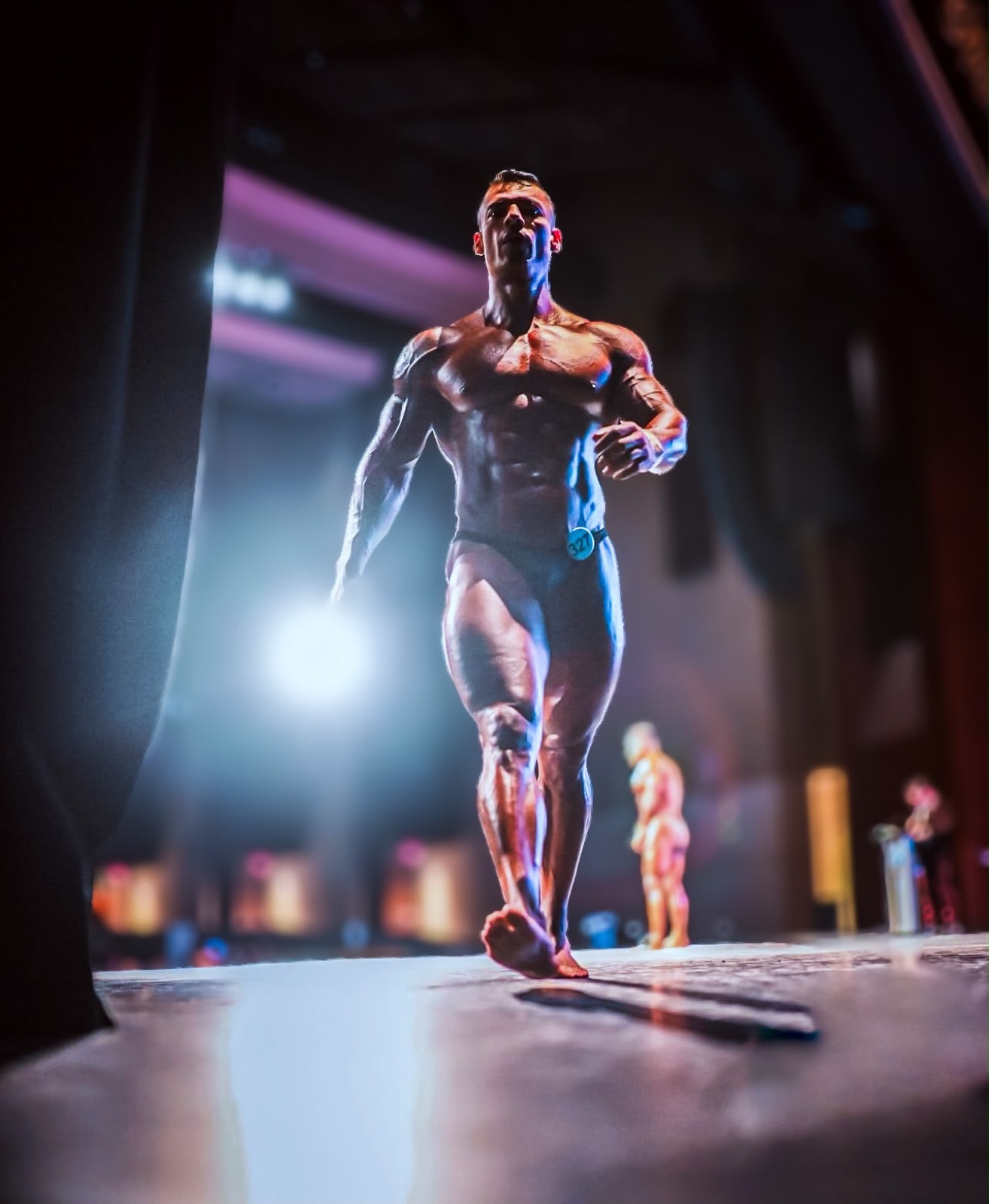 Gio Melchor walking onto the bodybuilding stage under spotlight
